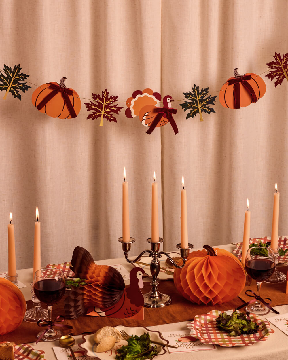 A cozy autumn-themed table setting with a paper honeycomb pumpkin, a turkey-shaped paper lantern, a pumpkin garland with leaves, lit taper candles, and fall dishes in front of a beige curtain backdrop.