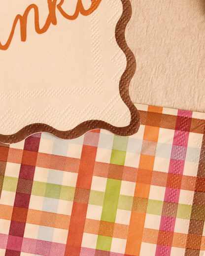 A close-up of a decorative paper napkin with a scalloped brown border and a multicolor plaid pattern in orange, pink, green, and blue on a cream background.