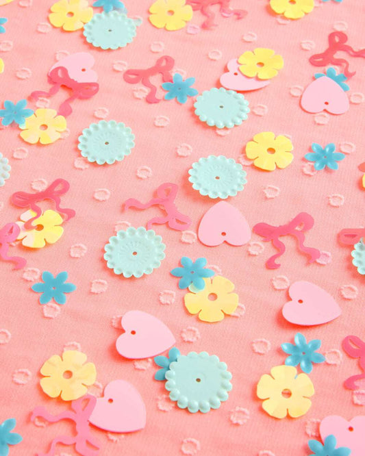 Pink, blue, and yellow plastic confetti in flower, heart, and bow shapes scattered on pink textured fabric.