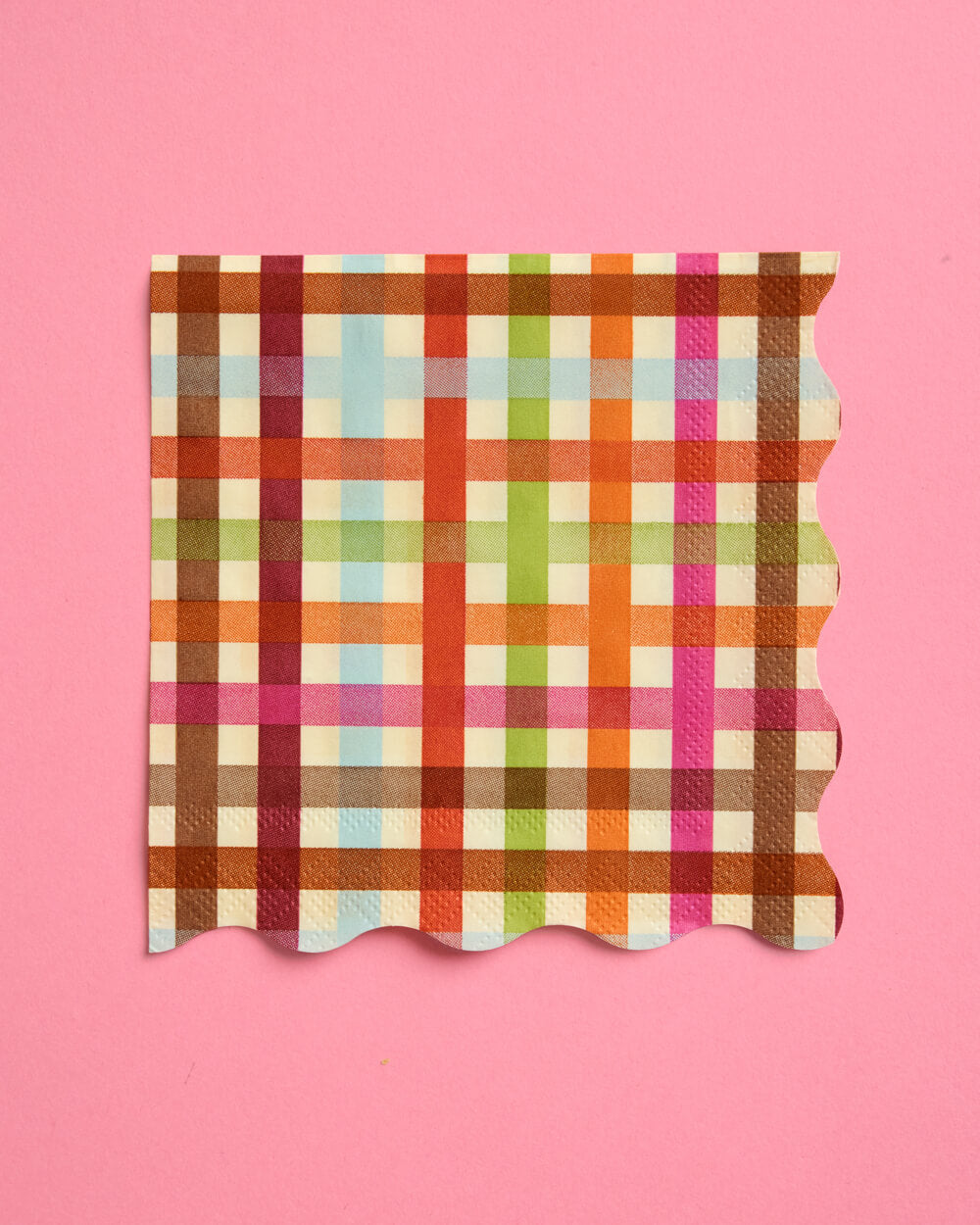 A square paper napkin with a colorful plaid pattern, featuring scalloped edges, displayed on a solid pink background.
