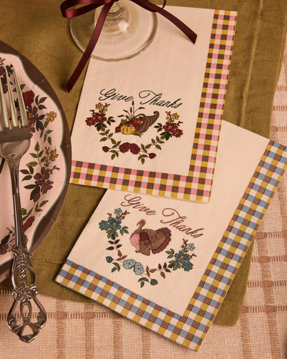 Two cream-colored paper napkins with a gingham border and a central 'Give Thanks' turkey and floral illustration, styled on a table with a plate, fork, and wine glass.
