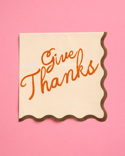 A square cream paper napkin with scalloped edges and the words 'Give Thanks' written in orange script, placed on a solid pink background.