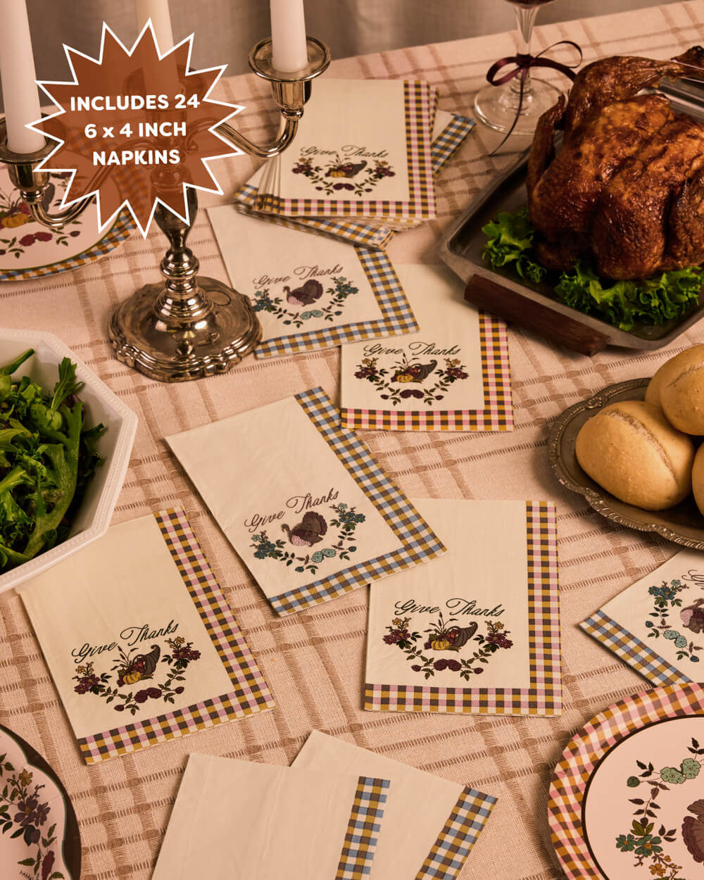 A styled Thanksgiving table setting showing multiple cream napkins with a rustic gingham border and a printed 'Give Thanks' chicken and acorn motif. The scene includes a roasted turkey, green beans, rolls, plates, and candlesticks on a checkered tablecloth, with a starburst callout stating 'INCLUDES 24 6 x 4 INCH NAPKINS'.