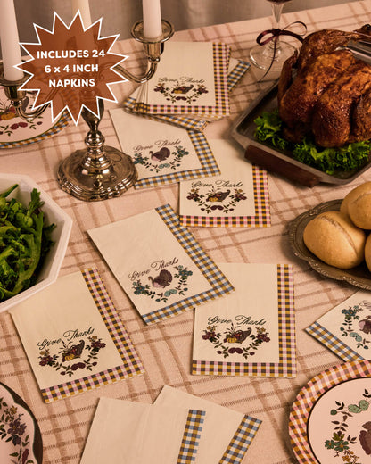 A styled Thanksgiving table setting showing multiple cream napkins with a rustic gingham border and a printed 'Give Thanks' chicken and acorn motif. The scene includes a roasted turkey, green beans, rolls, plates, and candlesticks on a checkered tablecloth, with a starburst callout stating 'INCLUDES 24 6 x 4 INCH NAPKINS'.