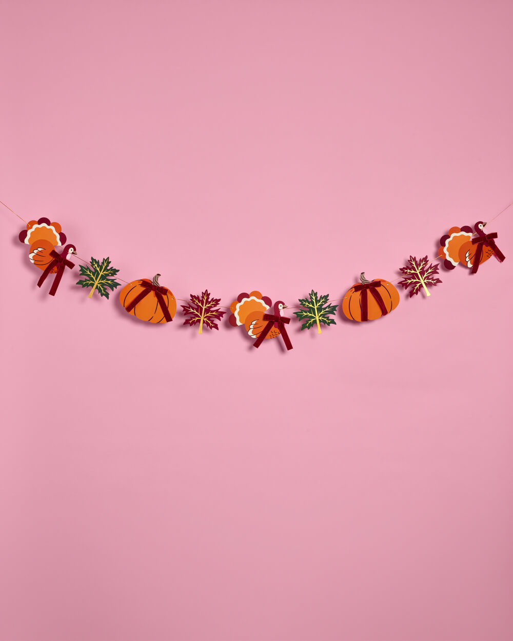 A festive autumn-themed paper garland featuring orange pumpkins, candy corn, maple leaves, and ribbons, strung across a solid pink background.