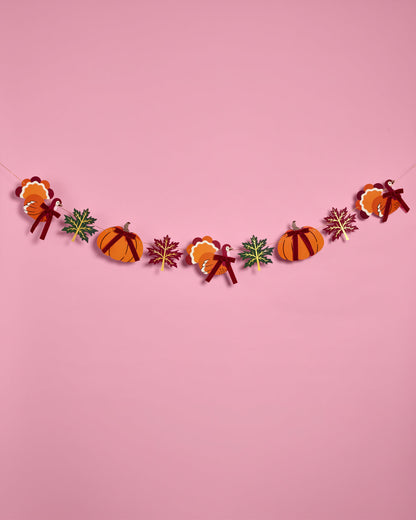 A festive autumn-themed paper garland featuring orange pumpkins, candy corn, maple leaves, and ribbons, strung across a solid pink background.