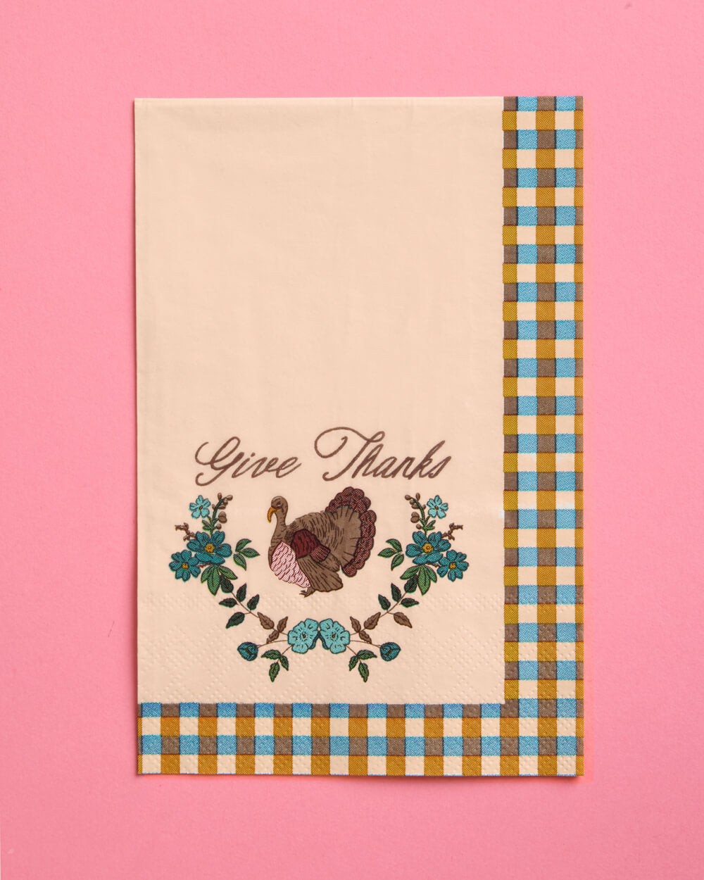 A square cream-colored fabric napkin with a gingham border in blue, yellow, and white, featuring an embroidered turkey and floral wreath with the cursive text 'Give Thanks' on a solid pink background.