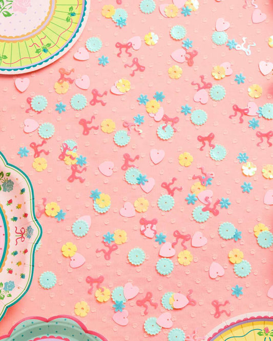 Pastel pink, blue, and yellow plastic confetti in heart, flower, and bow shapes scattered on a pink surface.