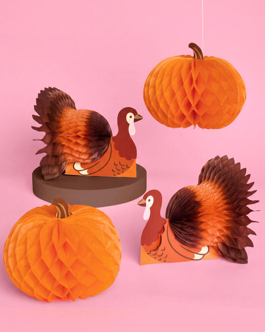 Paper honeycomb Thanksgiving party decorations featuring orange pumpkins and brown turkeys on a pink background.