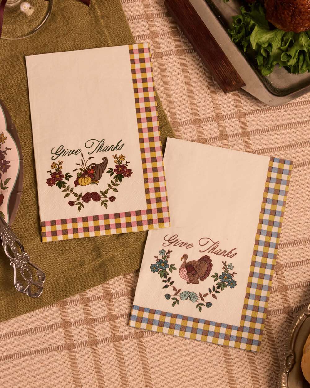 Two decorative paper napkins with a Thanksgiving theme, featuring 'Give Thanks' text, floral and bird illustrations, and gingham borders, styled on a table with a green placemat and checkered tablecloth.