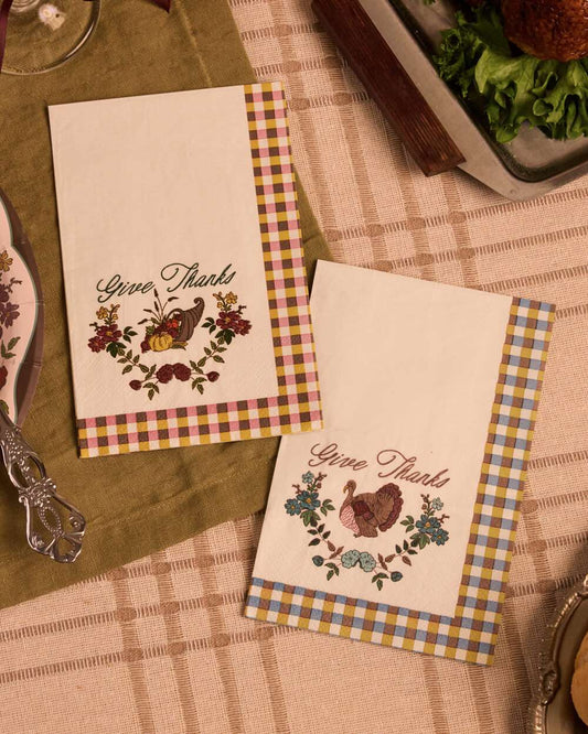 Two decorative paper napkins with a Thanksgiving theme, featuring 'Give Thanks' text, floral and bird illustrations, and gingham borders, styled on a table with a green placemat and checkered tablecloth.