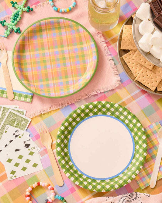 Two round paper plates with green gingham and plaid patterns on a colorful tablecloth with playing cards and wooden forks.