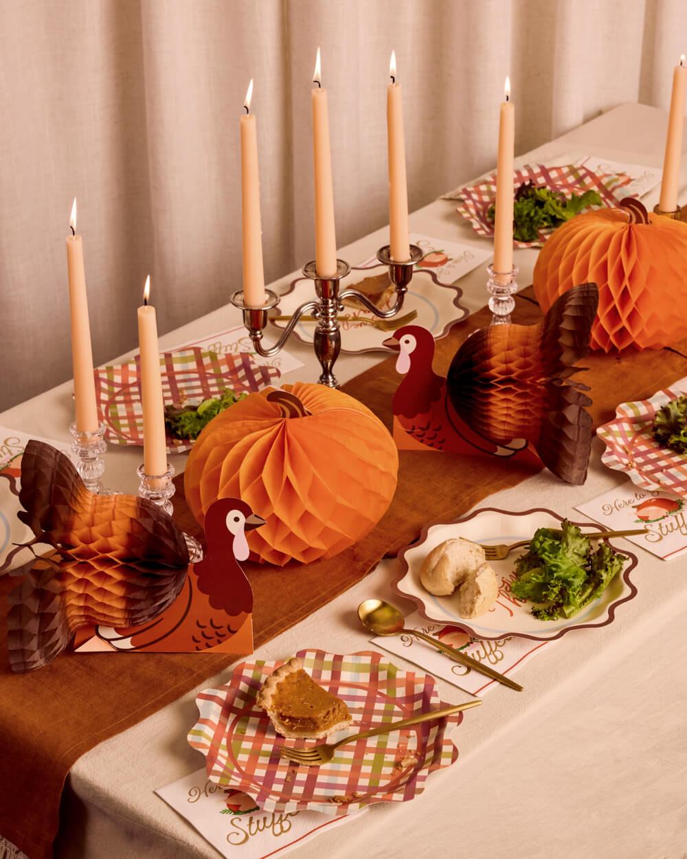 A styled Thanksgiving table setting with orange honeycomb paper decorations, turkey cutouts, lit taper candles, checkered place settings, and serving dishes on a draped tablecloth.