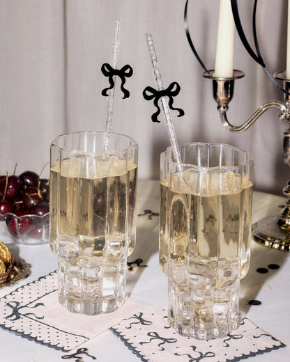 Two clear faceted drinking glasses filled with a pale golden beverage, set on a table with decorative napkins, cherries, a candle holder, and bow-shaped straws.