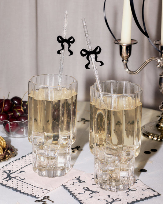 Two clear faceted drinking glasses filled with a pale golden beverage, set on a table with decorative napkins, cherries, a candle holder, and bow-shaped straws.