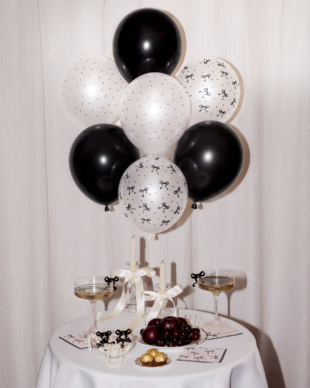 Black and white balloon bouquet decoration with black and white balloons, styled on a white tablecloth with champagne glasses, candles, and fruit.