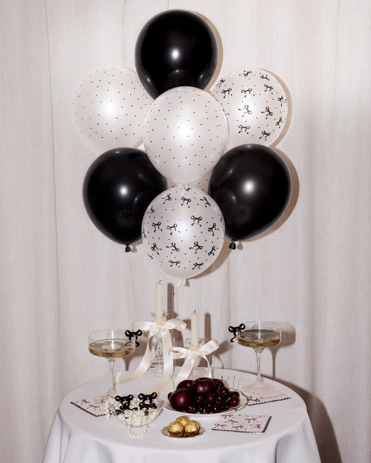 Black and white balloon bouquet decoration with black and white balloons, styled on a white tablecloth with champagne glasses, candles, and fruit.