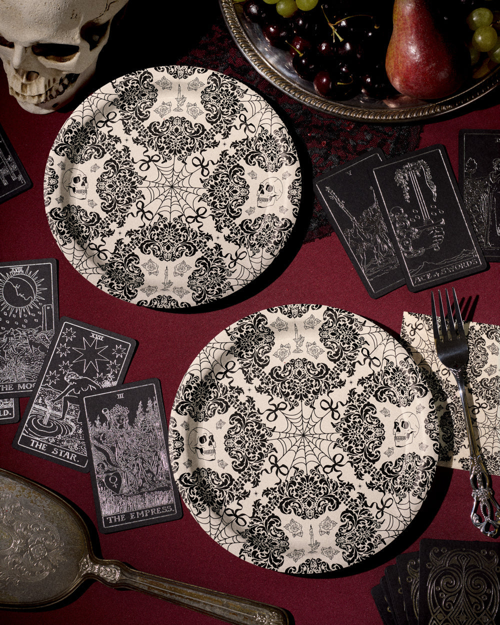 Decorative black and white plates with skull and spider web patterns on a red tablecloth with tarot cards and a skull.