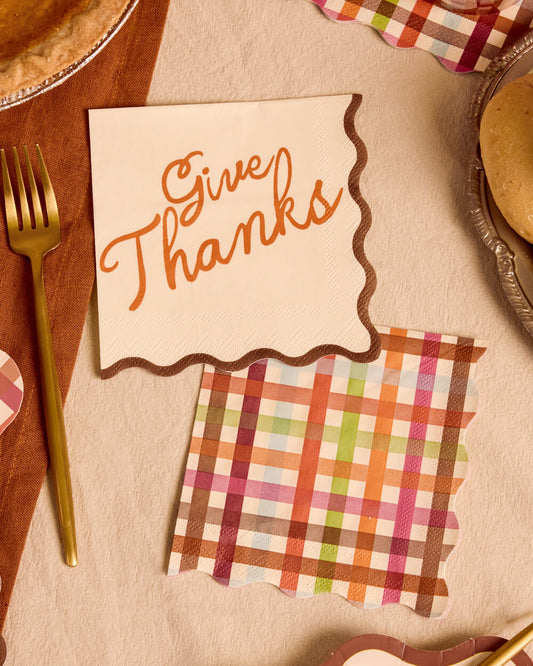 Thanksgiving-themed paper napkins on a beige tablecloth, including one with 'Give Thanks' in orange script and one with a multicolor plaid pattern, styled with a gold fork and pie dishes.