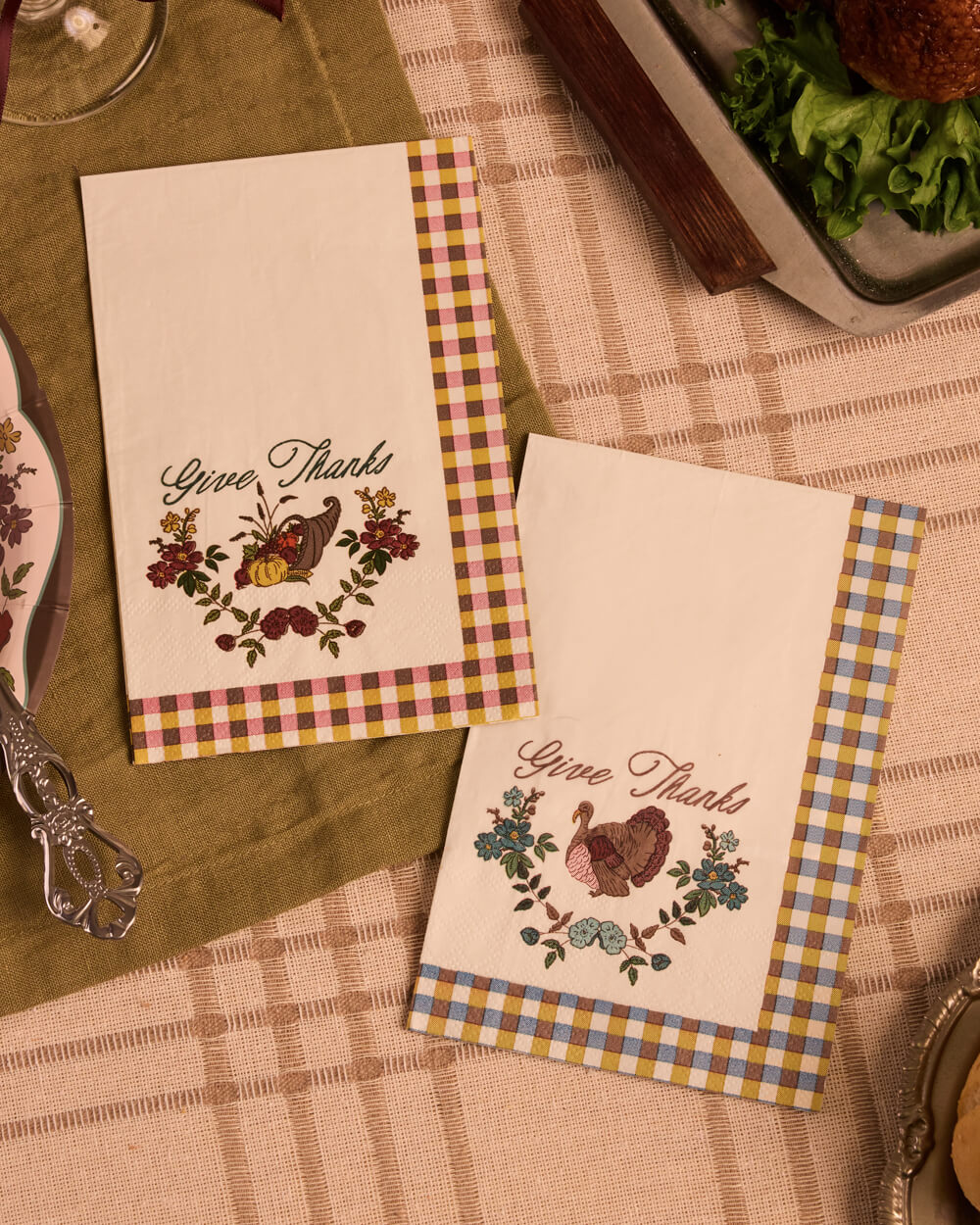 Two decorative paper napkins with a Thanksgiving theme, featuring 'Give Thanks' text, floral and bird illustrations, and gingham borders, styled on a table with a green placemat and checkered tablecloth.