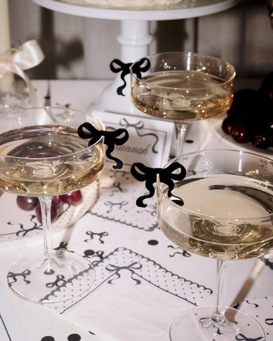 Three stemmed wine glasses filled with white wine, each decorated with a small black bow-shaped charm, arranged on a patterned table runner with grapes and a cake stand in the background.