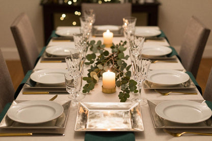 Holiday tablescape featuring silver square charger plates under white china with gold flatware and simple candles and greenery.