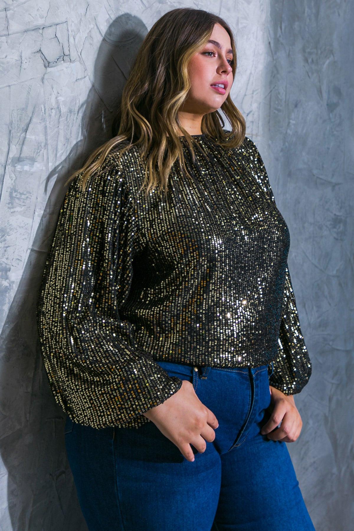Woman wearing a gold sequin top and blue jeans against a textured gray wall.