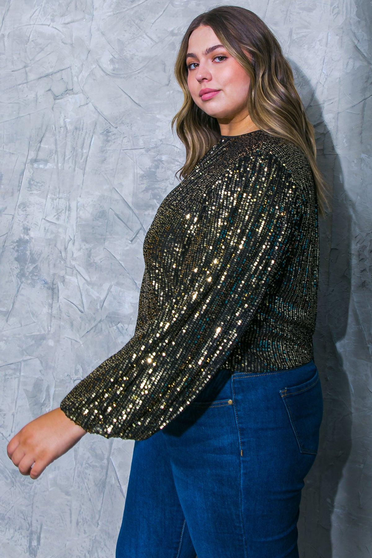Woman wearing a gold sequin top and blue jeans against a textured gray wall.