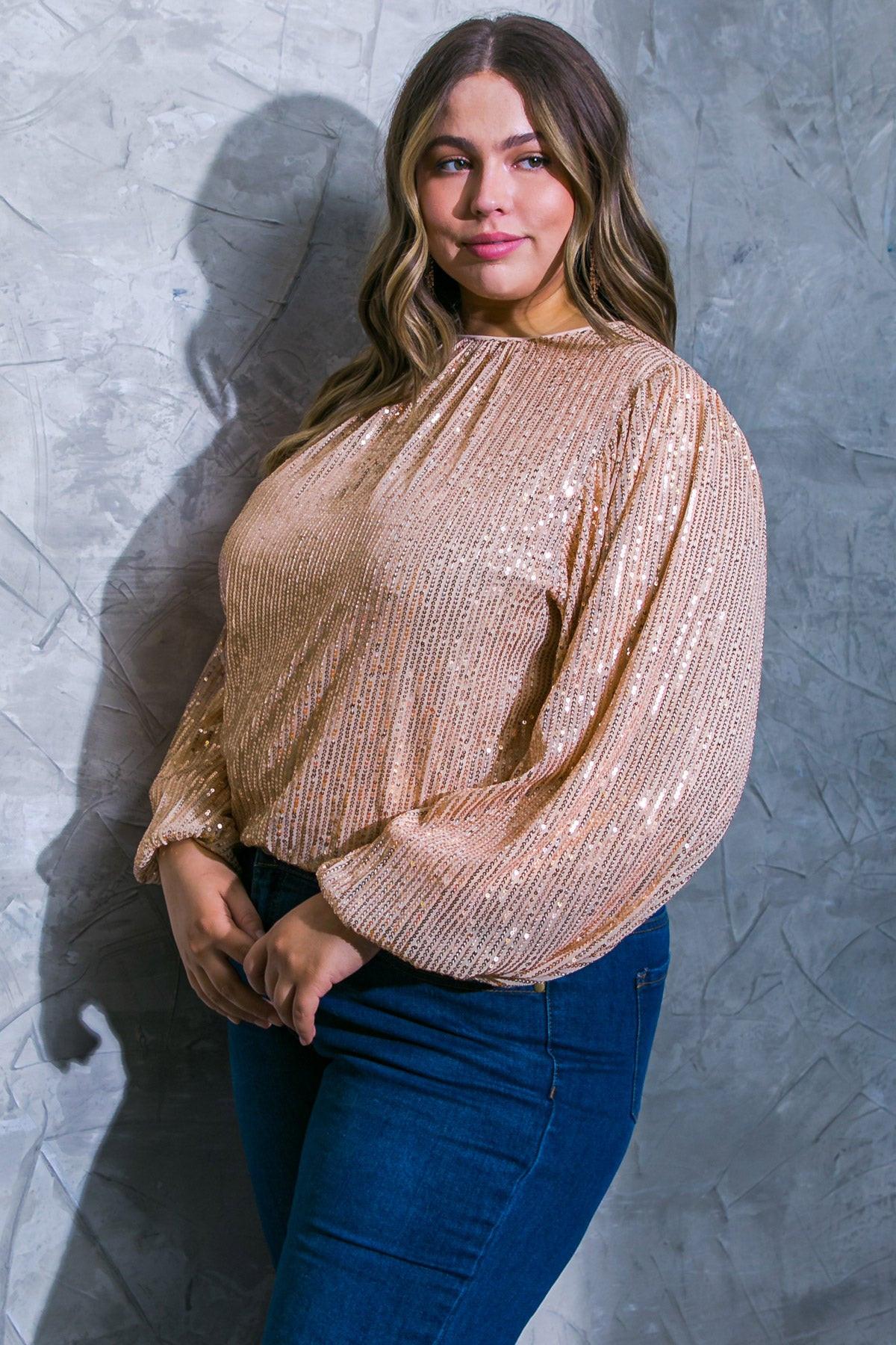 Woman wearing a sparkly beige top and blue jeans against a textured gray wall.
