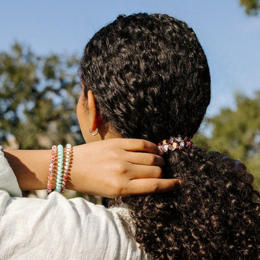 Three patterned and teal plastic spiral hair ties worn on a wrist and one securing a low ponytail in dark curly hair.