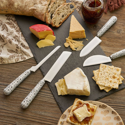 Tile-patterned ceramic handle cheese knives arranged with cheeses, bread, crackers, and jam on a slate board.