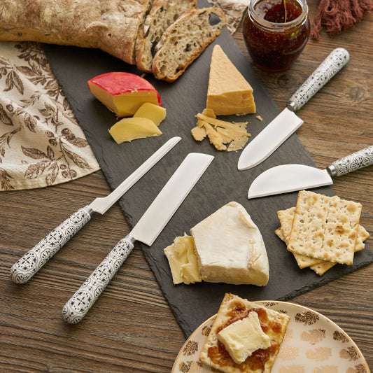 Tile-patterned ceramic handle cheese knives arranged with cheeses, bread, crackers, and jam on a slate board.