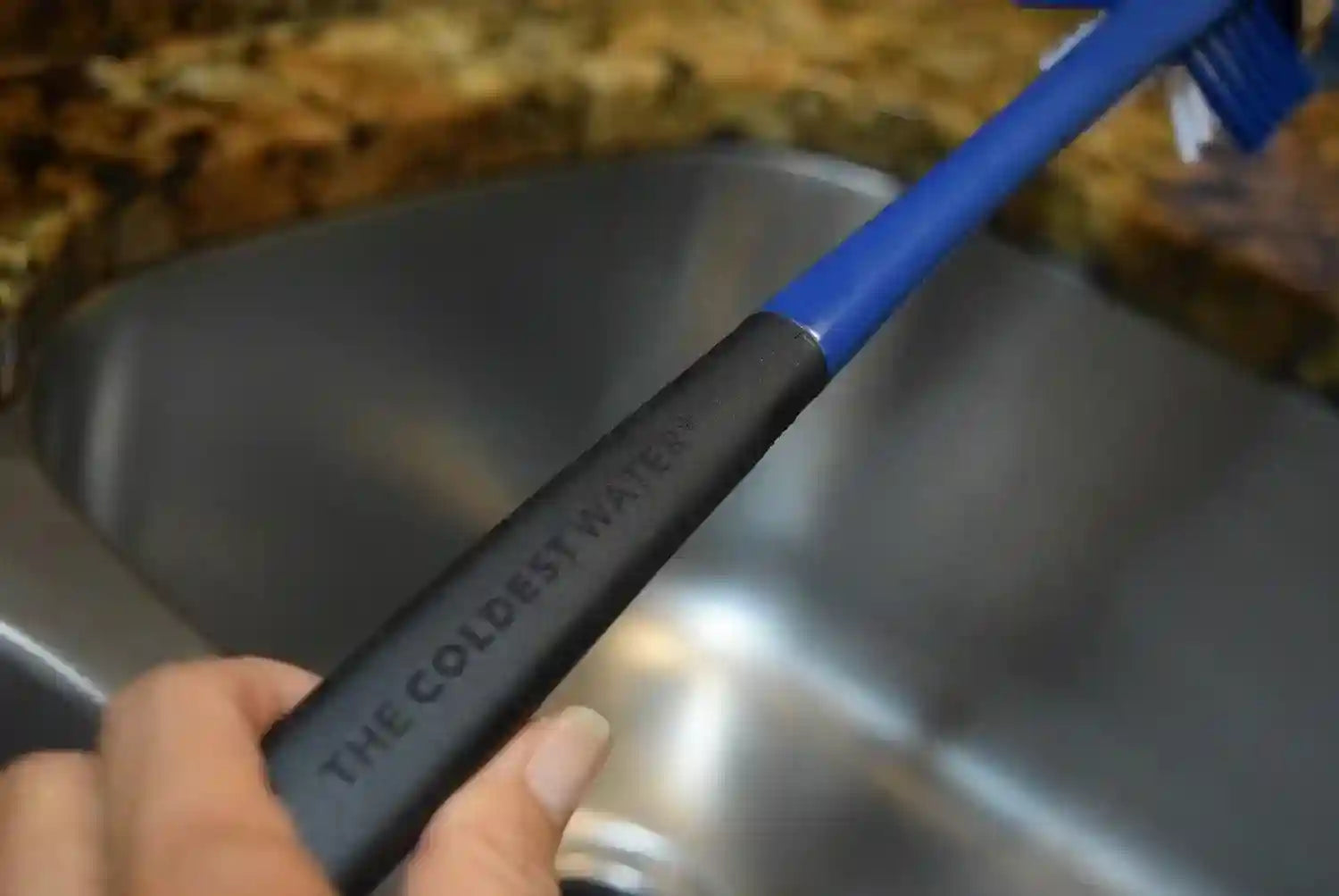 Blue and black scrub brush with 'The Coldest Water' branding held over a sink.