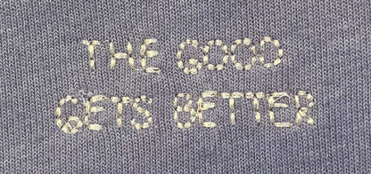 Close-up of a gray knit fabric with cream-colored embroidered text reading 'THE GOOD GETS BETTER'.