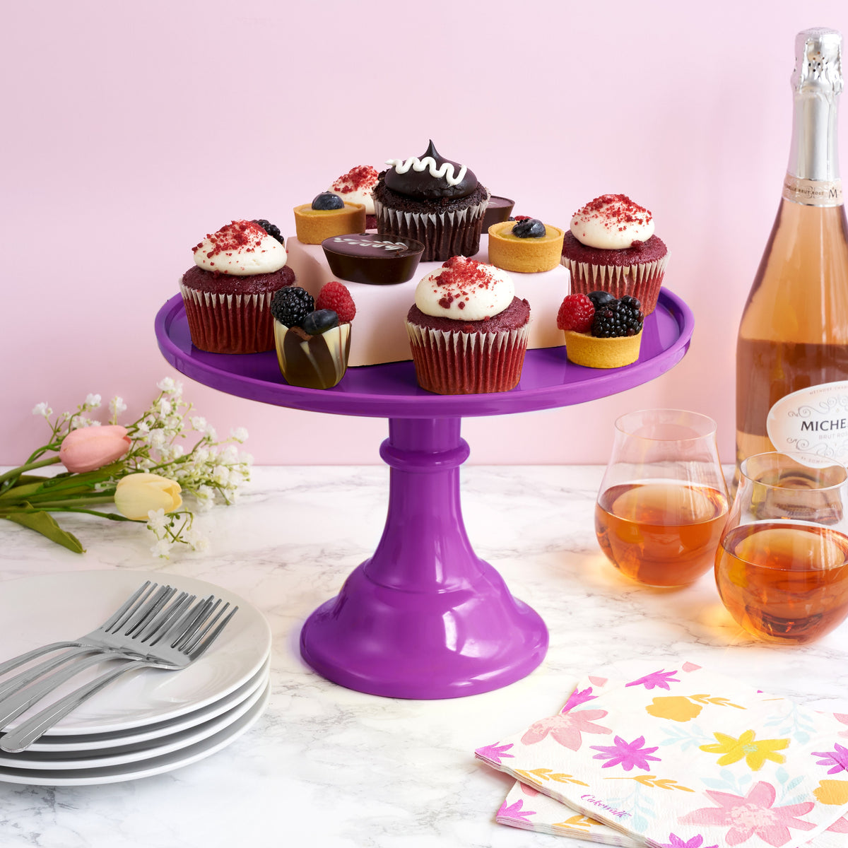 Purple cake stand with cupcakes and a bottle of champagne on a table with glasses and flowers.