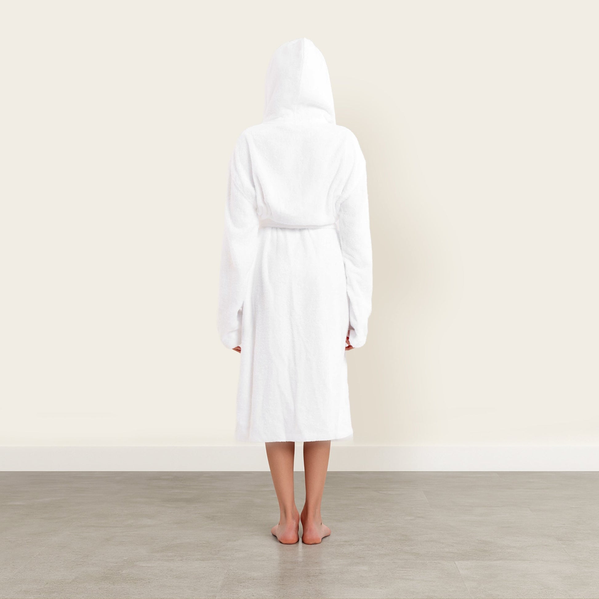 Person wearing a white robe standing in a minimalistic room with a light-colored wall and wooden floor.