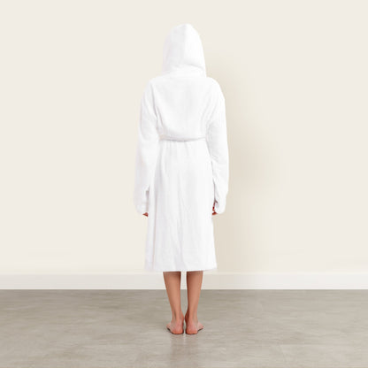 Person wearing a white robe standing in a minimalistic room with a light-colored wall and wooden floor.