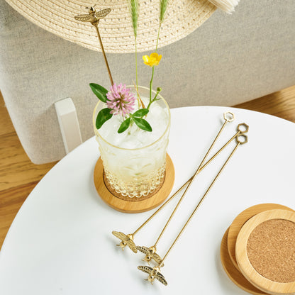 Gold bumble bee stir sticks styled with a flower-garnished iced drink on a white table.