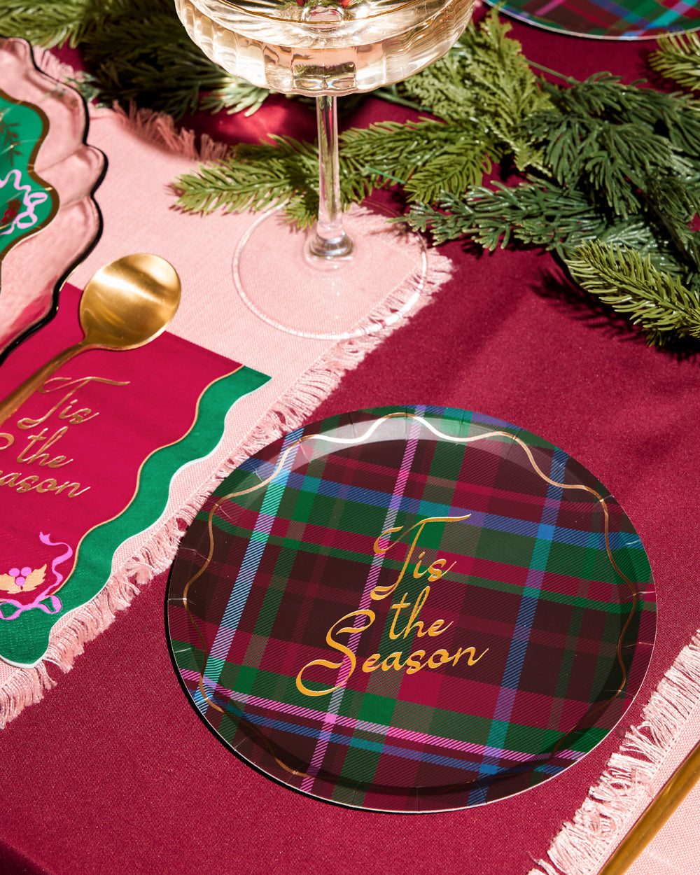 Plaid plate with 'Tis the Season' text on a festive table setting with a glass of wine and greenery.