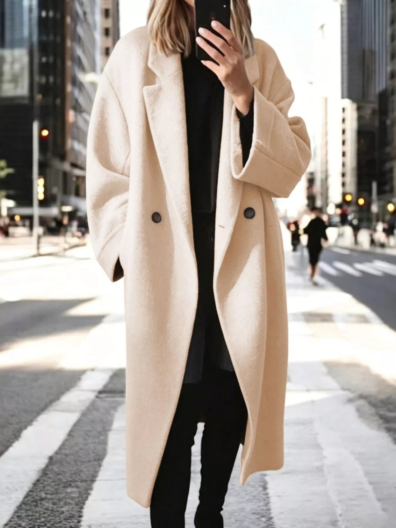 A woman wearing a long, oversized cream-colored wool coat over a black outfit, standing on a city crosswalk with blurred skyscrapers in the background.