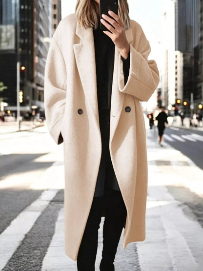 A woman wearing a long, oversized cream-colored wool coat over a black outfit, standing on a city crosswalk with blurred skyscrapers in the background.