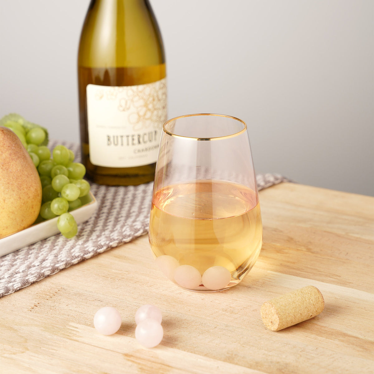 Rose quartz wine gems in a gold-rimmed glass of white wine, with extra gems on a wooden table.