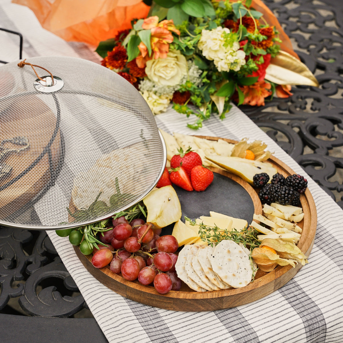 Wooden platter with assorted fruits, cheeses, and crackers on a checkered tablecloth with a floral arrangement in the background.