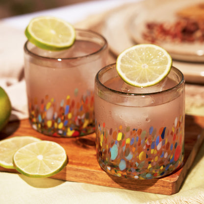 Two colorful speckled Artistico tumblers filled with iced pink drinks and lime slices on a wooden board.
