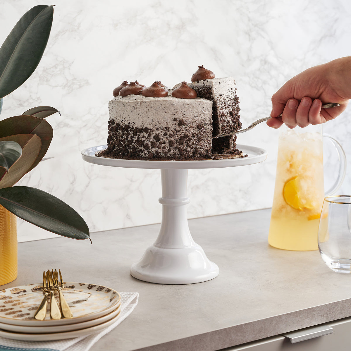 Chocolate cake on a white stand with a hand cutting a slice, surrounded by plates and a pitcher of lemonade.