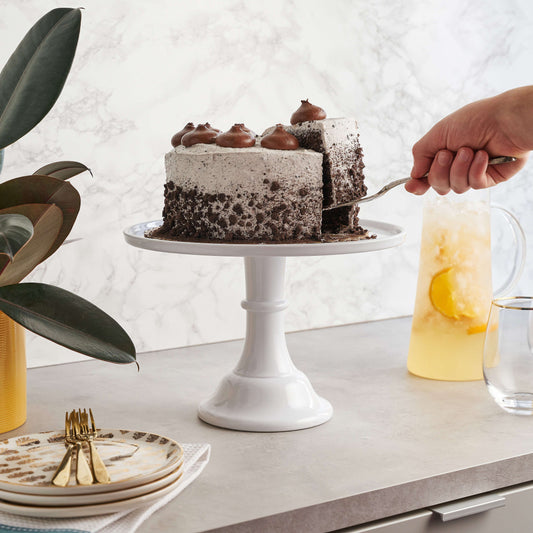 Chocolate cake on a white stand with a hand cutting a slice, surrounded by plates and a pitcher of lemonade.